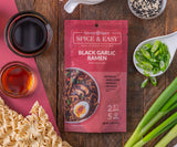 Black Garlic Ramen Spice & Easy package, top down with green onions, soy sauce, scallions, and ramen noodles arranged around it on a wood table.