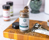 Jar of Pikes Peak Butcher's Rub on a wood cutting board with a silver measuring spoon taking spices out of the jar. 